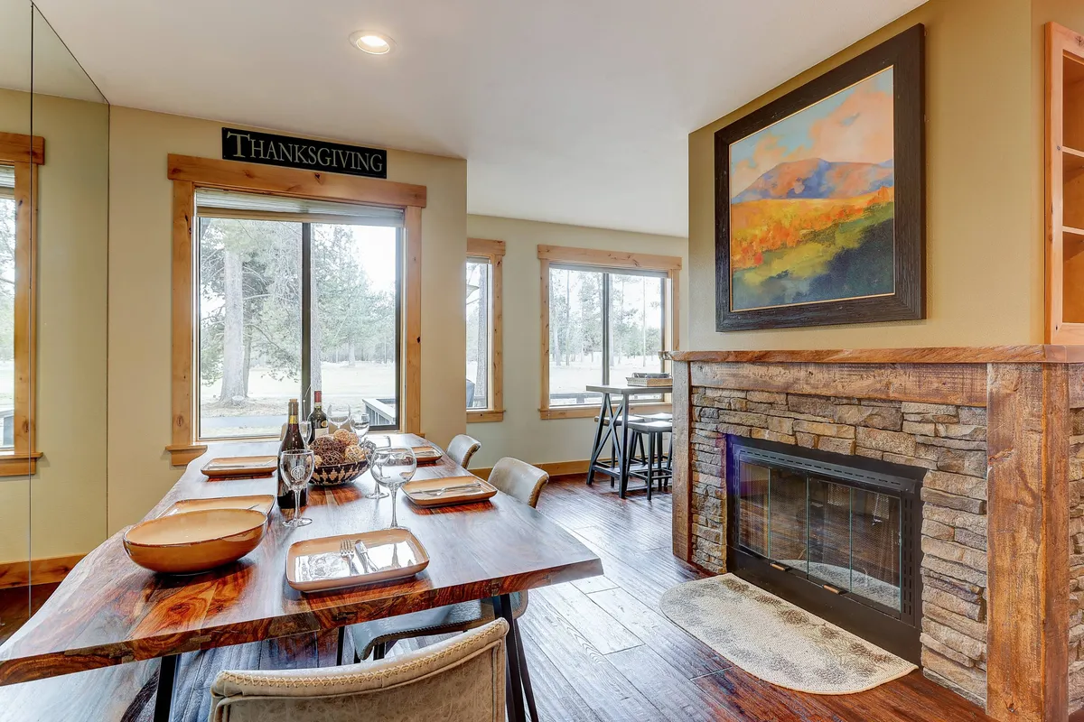 A cozy dining area with a wooden table set before a fireplace, surrounded by artwork and windows that stream in sunlight. The setting invites family gatherings, festive meals, and intimate breakfasts, perfectly embodying the social heart of the home.