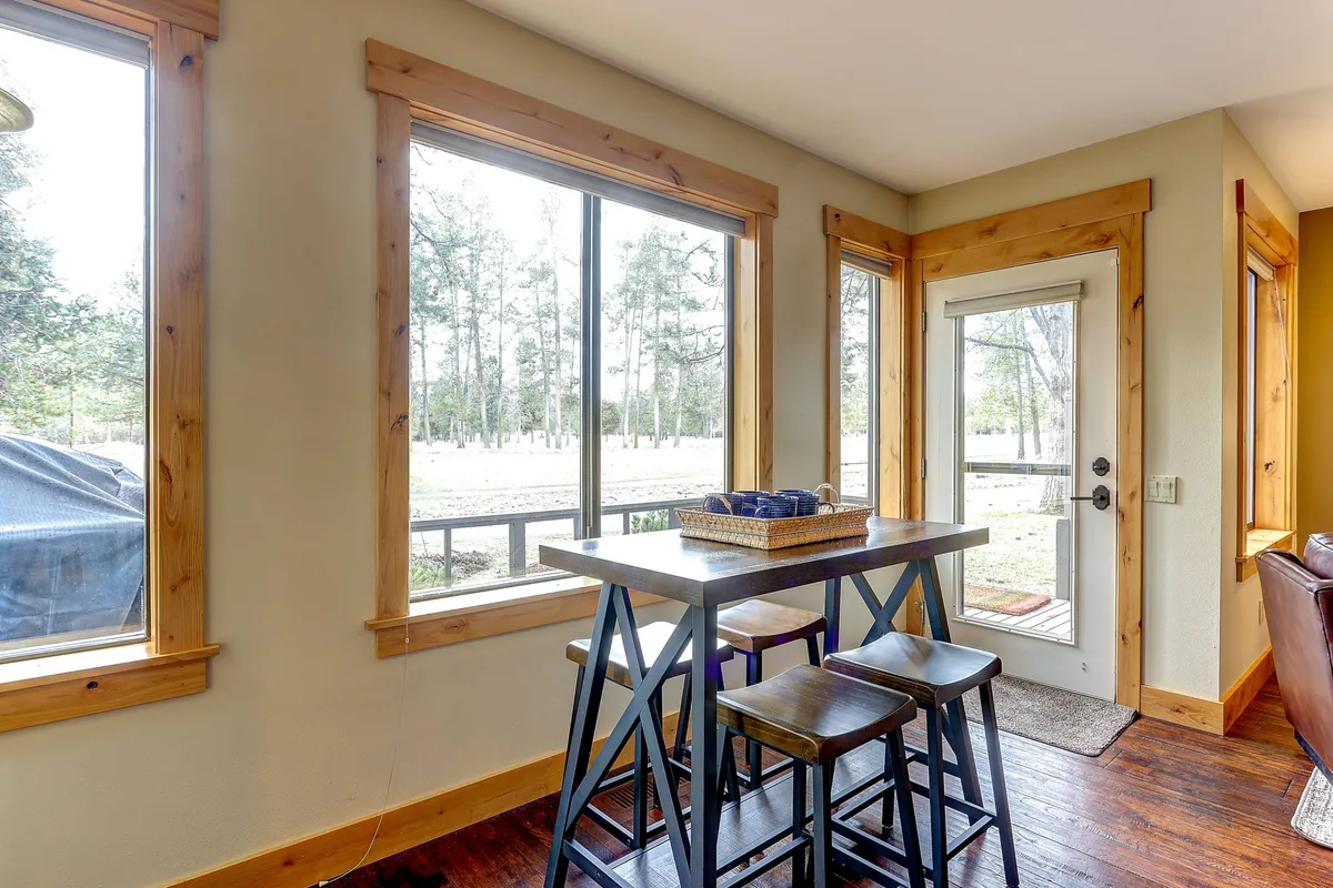 A dining space flooded with natural light from large windows, featuring a wooden table and stools, and a door leading outside. The room seamlessly blends the indoors with the outdoors, suggesting easy access to fresh air, patios, or backyard areas.
