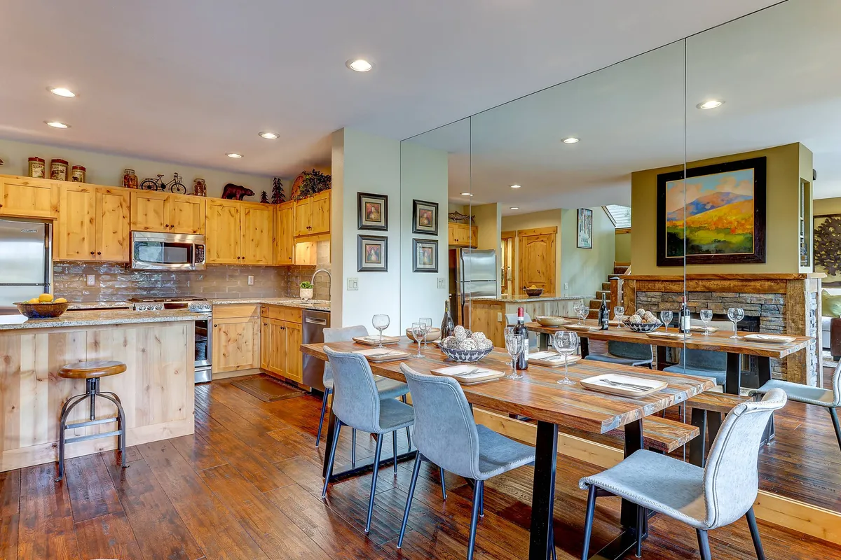 The image showcases a warm, sunlit dining area centered around a beautiful wooden table, with a stylish kitchen island in the background. A mirrored wall amplifies the airy feel, while tasteful artwork adds character, creating an inviting space ideal for gathering, sharing meals, and making memories.