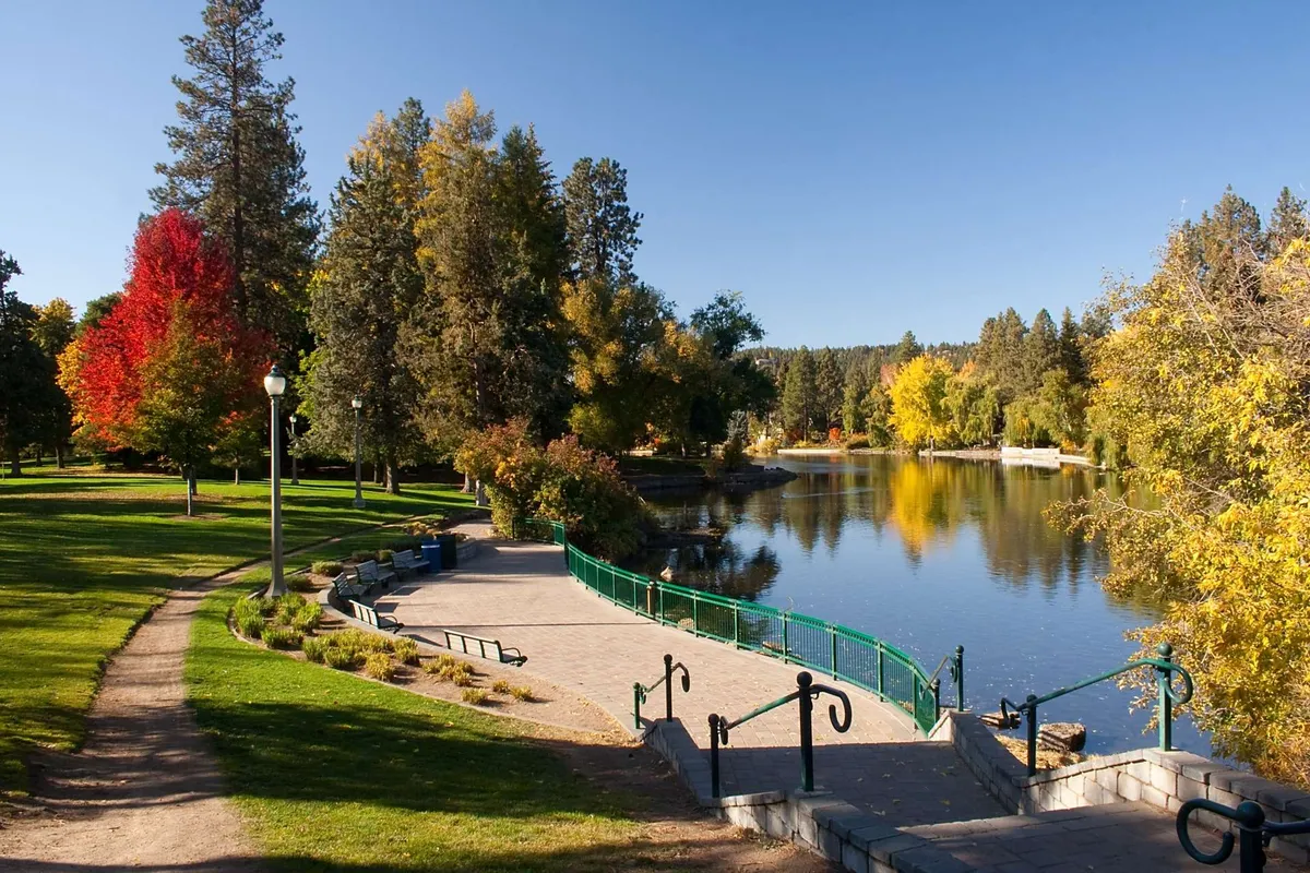 A peaceful lake scene bordered by colorful autumn trees, with a walking path and green railing inviting exploration. The area suggests opportunities for walking, fishing, and enjoying natural beauty year-round.