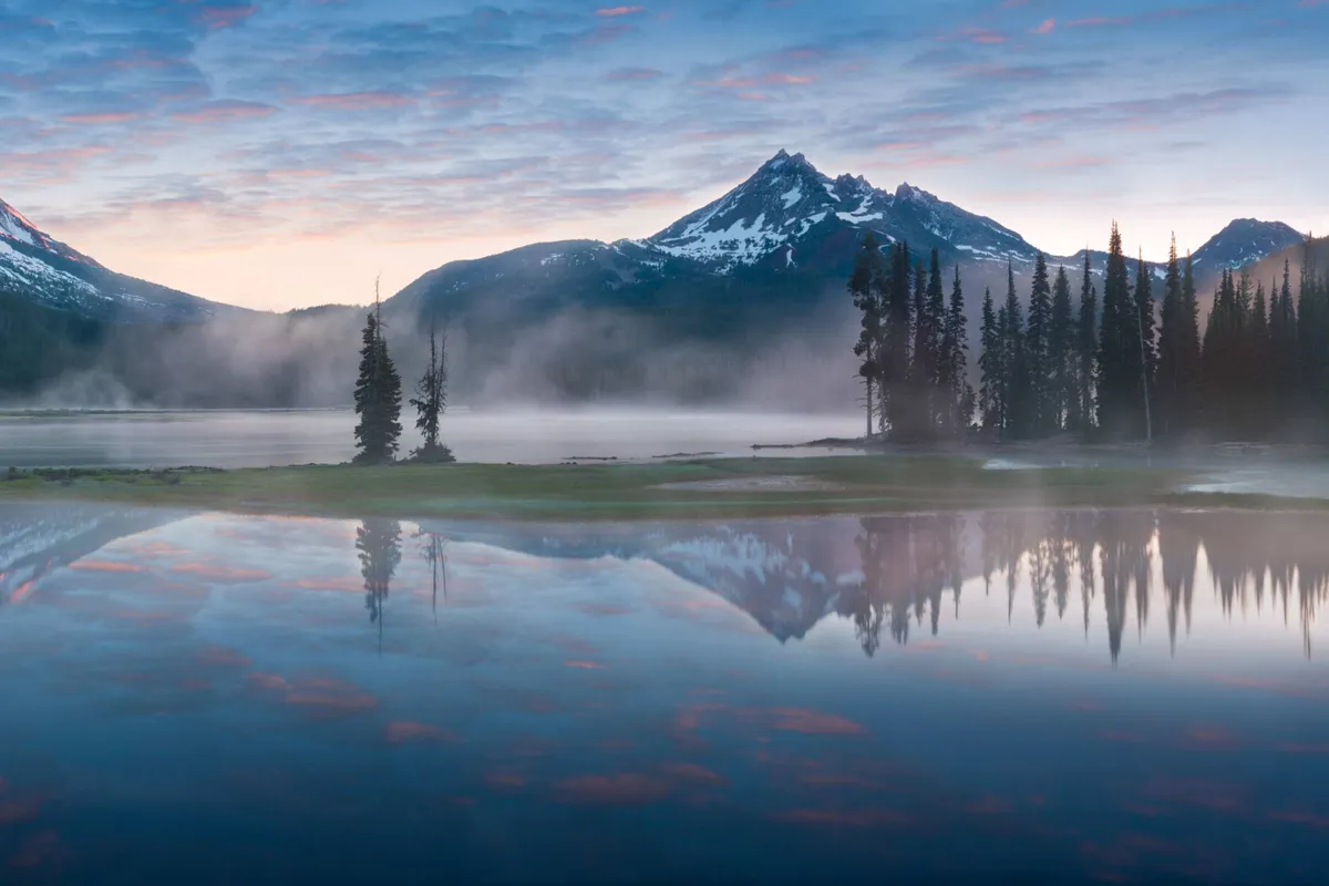 A tranquil scene showcasing misty mountains reflected on calm water, bordered by trees. The natural beauty of the landscape evokes a sense of outdoor adventure and exploration in the Sunriver area, making it ideal for activities like hiking with a dog or enjoying nature with a pet.