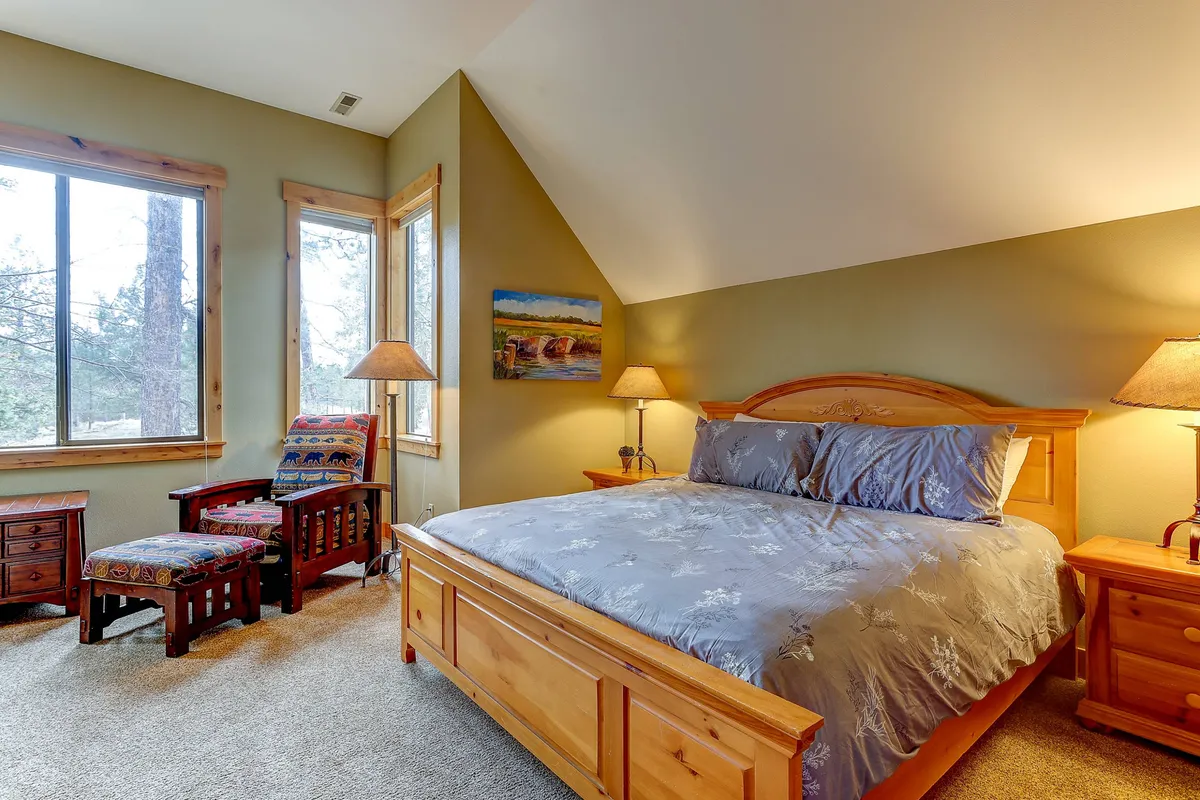 A serene bedroom featuring a queen-sized bed with gray bedding, wooden bedside tables, a large window allowing natural light to flood in, painted landscape artwork, and comfortable chairs for lounging.