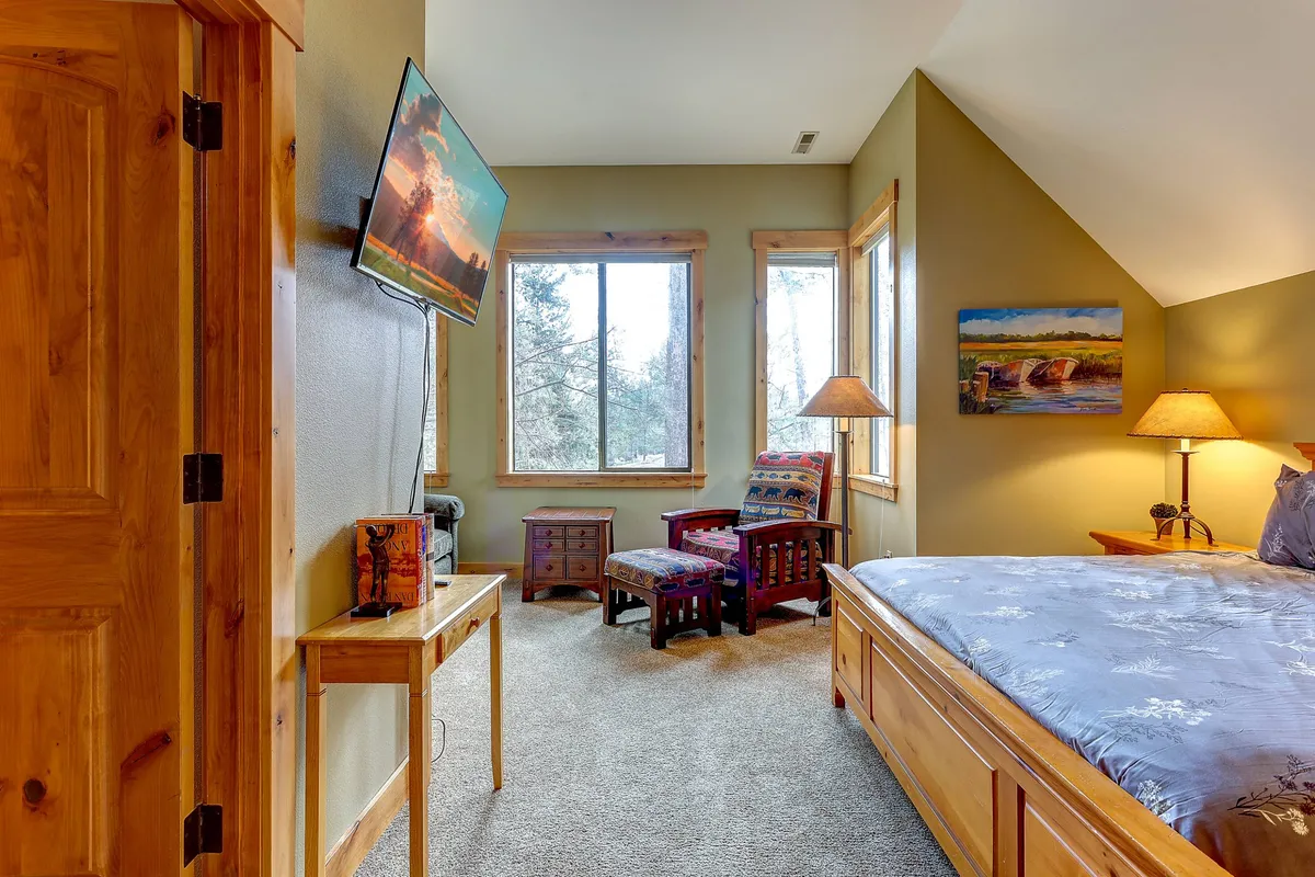 A cozy bedroom with a comfortable bed, television, large windows, an armchair, and wooden furniture. The room is bathed in natural light and offers serene views, perfect for relaxing or enjoying quiet moments.