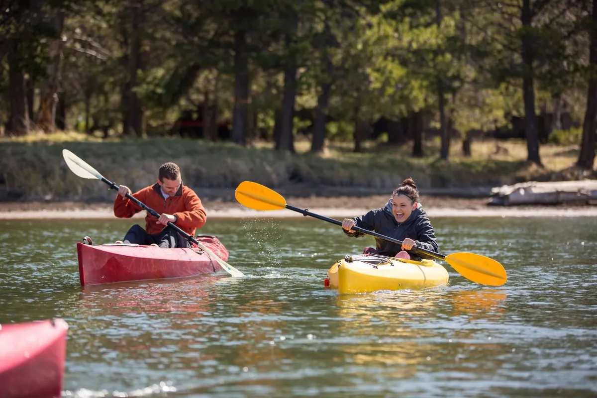 Two kayakers paddle brightly colored kayaks across a calm waterway, framed by trees and reflecting the energy and playfulness of outdoor recreation in Sunriver.