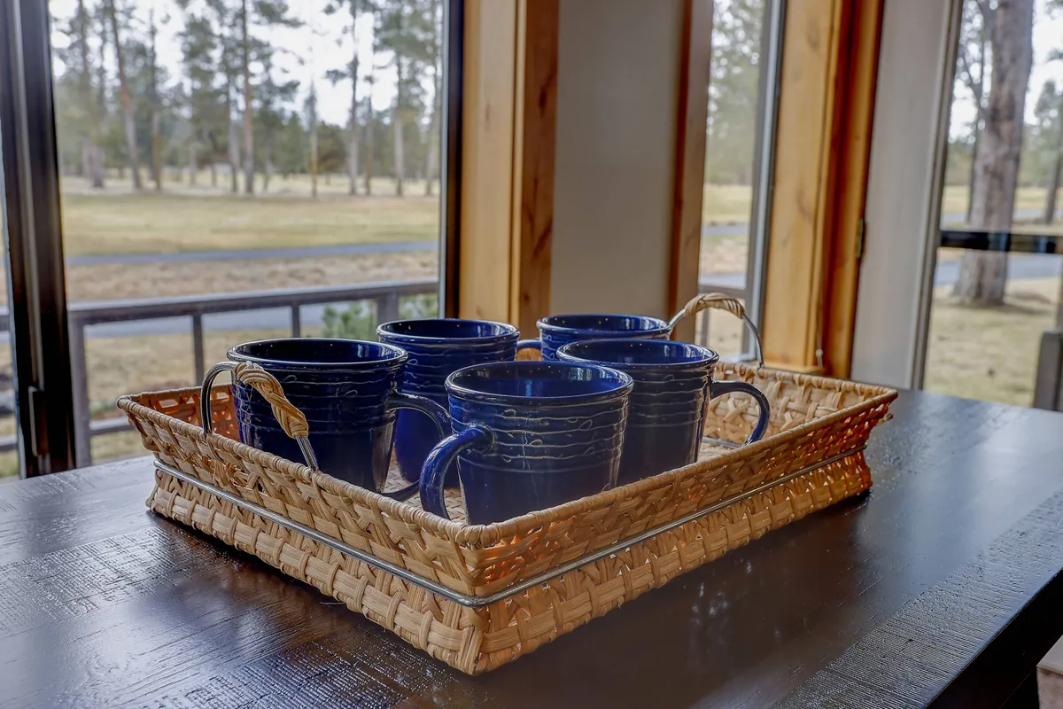 A cozy scene featuring blue mugs on a wooden table and a woven basket, set beside large windows that let in soft natural light. The setup suggests a quiet morning coffee or intimate conversation while enjoying the view outside.
