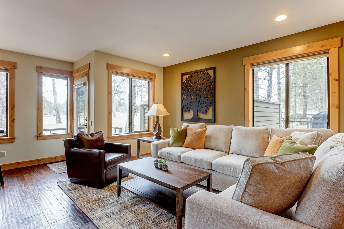 A bright and welcoming living room featuring a spacious sectional sofa, wooden coffee table, and decorative wall art. Large windows let in abundant natural light and offer scenic views, creating a relaxing and inviting atmosphere ideal for gatherings and quiet moments alike.