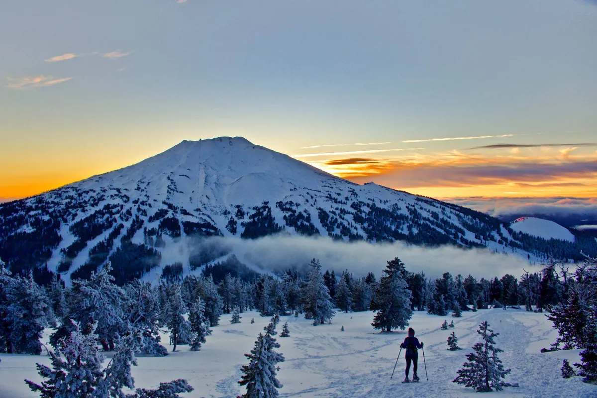 A striking view of a snow-covered mountain framed by tall pine trees, with a skier in action and a vibrant sunset sky overhead. The landscape exudes adventure and natural beauty, hinting at thrilling winter sports and year-round exploration possibilities.
