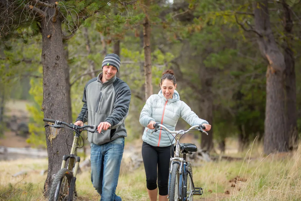 This image captures two cyclists walking their bikes along a scenic pathway surrounded by tall trees. The sunlight filters through the foliage, creating a warm and inviting atmosphere that highlights the outdoor recreational opportunities in Sunriver.