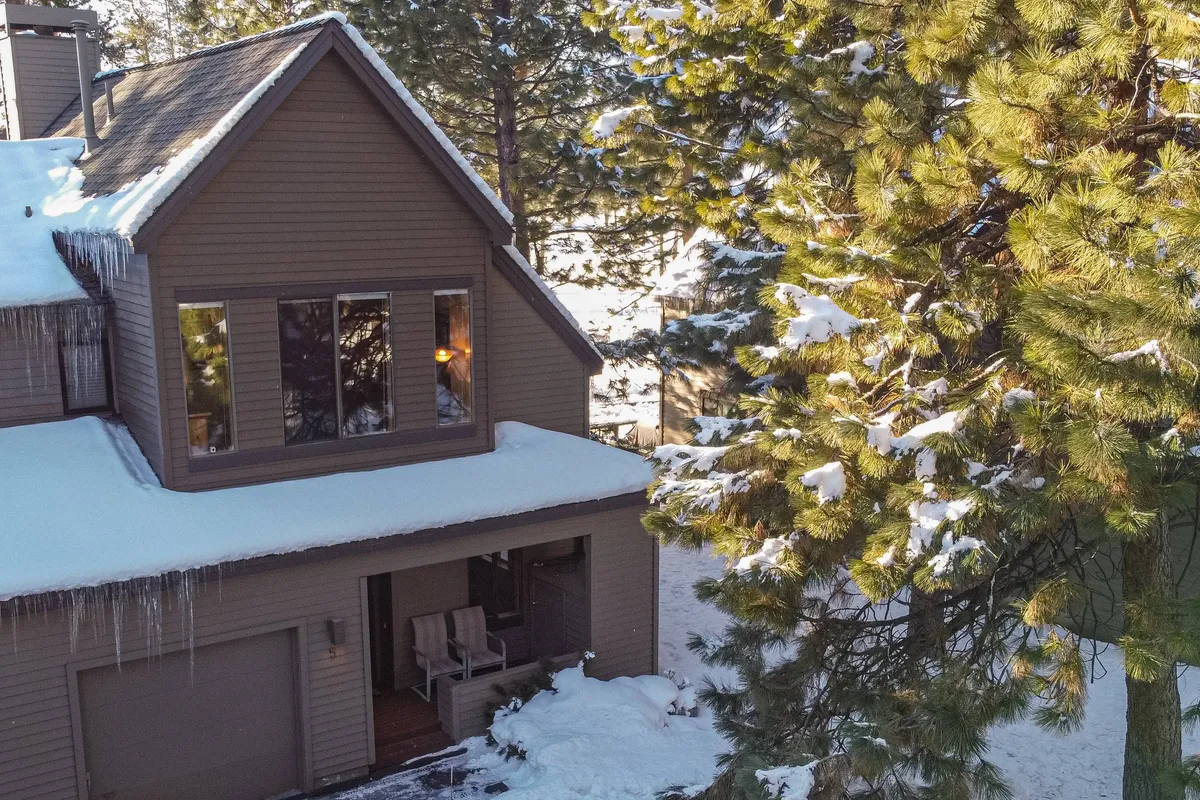 A welcoming view of the house nestled among snow-covered trees, featuring a front porch with chairs perfect for relaxing after outdoor activities. The setting naturally connects the home to its forested surroundings and trail access.